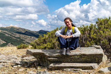 Dalgalı saçlı sarışın kadın, bir dağ yolunda dinlenirken bir kayanın üzerinde oturuyor..
