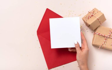 Kadın elinde boş kare kağıt maket zarf kırmızı, altın yıldızlı konfeti, bej arka planda hediye kutuları var. Düz konum, üst görünüm, kopyalama alanı, minimalist. Noel ve Yeni Yıl kavramı