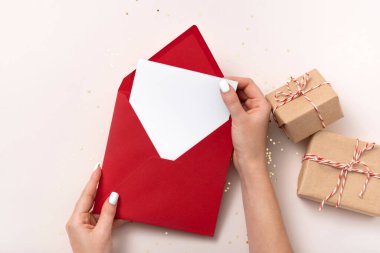 Kadın elinde boş kare kağıt maket zarf kırmızı, altın yıldızlı konfeti, bej arka planda hediye kutuları var. Düz konum, üst görünüm, kopyalama alanı, minimalist. Noel ve Yeni Yıl kavramı