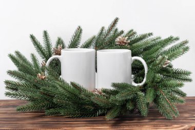 Kahverengi ahşap bir masada iki beyaz çay fincanı ve Noel ağacı çelengi dalları. Tasarımınız için kase şablonu, kopyalama alanı ile logo. Yeni Yıl