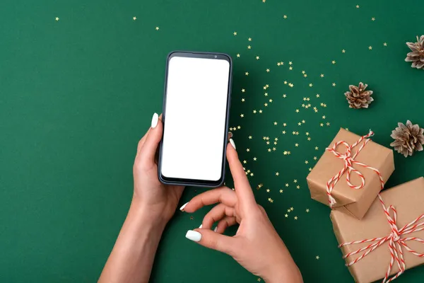 Çağdaş akıllı telefonu olan, yeşil arka planı olan ve Noel süslemeli kadın eller. Düz şablon. Firavun çelengi, hediye kutusu.