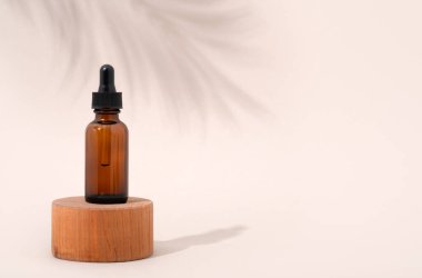 Mockup cosmetic face serum brown glass bottle with a pipette on a stylish minimalist beige white background wooden pedestal. Shadow from a palm tree branch. Female beauty concept, empty template