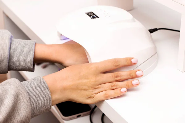 Woman puts hand with fresh beautiful pink modern gel polish manicure into led uv lamp for curing top cover of nailpolish. Process of drying gel on nails, close up. Beauty and self care