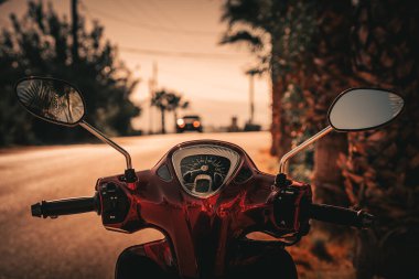 Kırmızı bir motosikletin ön tarafı bulanık bir yolun arka planında palmiye ağaçlarıyla kaplı..
