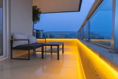 Large balcony of a luxury apartment. There is a small sofa with a coffee table. Evening time.