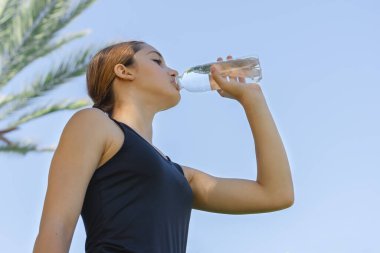 Mavi gökyüzüne karşı plastik şişeden su içen genç bir kızın düşük açılı görüntüsü.