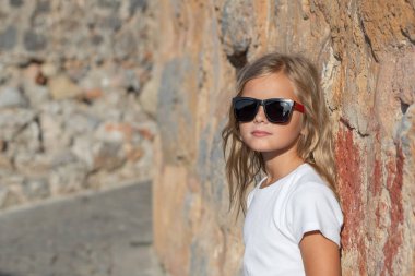 A young girl with blond hair and sunglasses leans her back against a stone wall and looks into the camera.