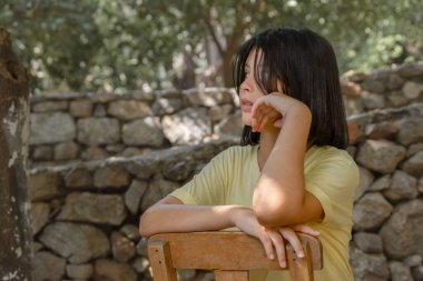 Genç bir kız tahta bir sandalyede oturur ve kameraya taş bir duvarın arkasında poz verir..