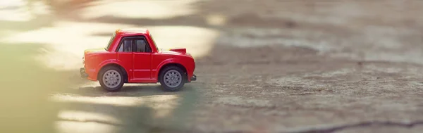 A model of a red toy car in the sunbeams on the road rides outdoors. Close-up. Horizontal format banner with place for text. Selective focus. Copy space — Stock Photo, Image