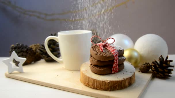 La farina bianca si versa sopra i biscotti natalizi legati con una corda rossa. Spuntino pomeridiano di anno nuovo con una tazza bianca e biscotti di gocce di cioccolato su uno stand di legno su un tavolo con pigne e palle. — Video Stock