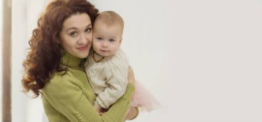 Süveter giyen genç bir anne bebeğe sarılıyor ve kameraya bakıyor. Pencerenin yanındaki gri arka planda kollarında bebek olan bir kadının portresi. Bakım, aşk, aile kavramı. Boşluğu kopyala