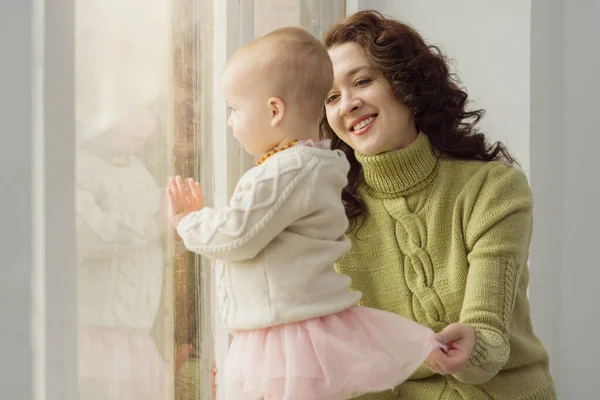 Das Porträt einer liebenden Mutter, die mit ihrem Baby spielt, während sie am Fenster sitzt. Eine schöne junge Frau bewundert das Baby, das aus dem Fenster schaut. Das Konzept von Familie, Fürsorge, Liebe, Aufrichtigkeit. — Stockfoto