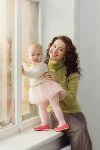 Una madre cariñosa con un bebé se sentará en la ventana. Mujer joven feliz con su hija en suéteres cálidos. Formato vertical, retrato. El concepto de cuidado, familia, felicidad. Copiar espacio —  Fotos de Stock
