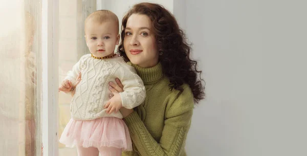 Horizontale Aufnahme der schönen jungen Mutter im Pullover mit ihrer kleinen Tochter, isoliert auf grauem Studiohintergrund. Porträt aus nächster Nähe, Blick in die Kamera. Kopierfläche für Werbung oder Promotion. — Stockfoto