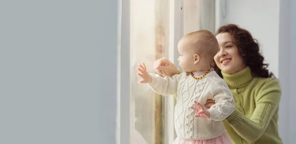 У окна сидит красивая молодая мама с ребенком в теплых свитерах. Понятие счастливой семьи, заботы, любви. Горизонтальный формат. Копирование пространства — стоковое фото