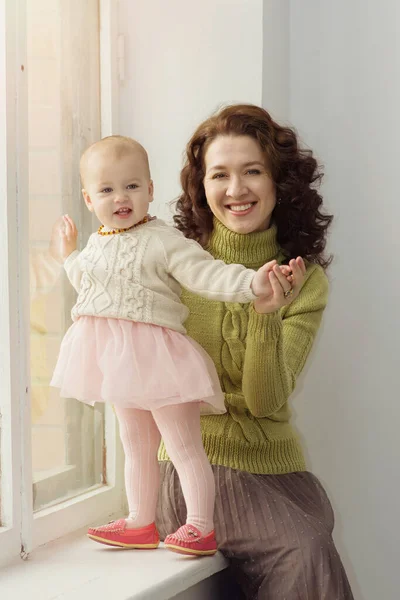 Красивая молодая мама с кудрявыми темными волосами в уютном вязаном свитере сидит у окна с маленькой девочкой на руках. Женщина с очаровательной дочерью смотрит в камеру и улыбается.. — стоковое фото