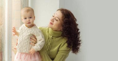 Pencerenin yanındaki gri arka planda, sıcak süveter giyen mutlu genç anne ve küçük kızı. Aile fotoğrafı. Mutlu aile kavramı, ilgi, sevgi. Yatay format. Boşluğu kopyala