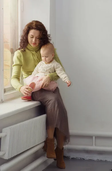 Una joven y hermosa madre sostiene al bebé en sus brazos y se pone los zapatos mientras está sentado en la ventana. La mujer está poniendo zapatos en su hija. El concepto de ayuda, cuidado, amor, familia. —  Fotos de Stock