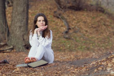 Beyaz elbiseli sakin kadın keskin tırnakları üzerinde durduktan sonra dinleniyor. Genç bir yogi kızı, sonbahar ormanında, dışarıda meditasyon yapmak için bir sadhu tahtasının yanında yerde oturuyor. Yakın plan. Boşluğu kopyala