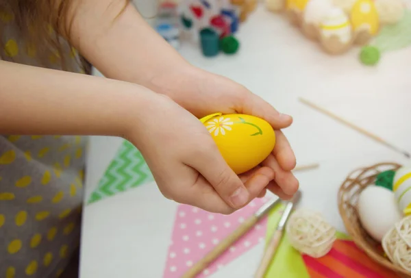 Dziecko trzyma w rękach żółte pisanki. Kreatywność z dziećmi. Dłonie dzieci z pisanką na tle białego stołu z farbami, pędzlami, jajkami, kolorowymi flagami wakacyjnymi. — Zdjęcie stockowe