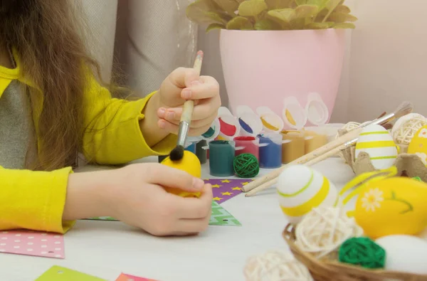Το παιδί βάφει πασχαλινά αυγά. Προετοιμασία για τις διακοπές του Πάσχα στο σπίτι. Η ευτυχισμένη κόρη κρατά ένα κίτρινο αυγό στα χέρια της. Κοντινό πλάνο παιδικών χεριών στο φόντο ενός λευκού ξύλινου τραπεζιού. Φωτογραφία Αρχείου