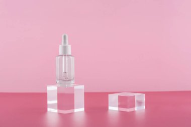Pembe arka planda akrilik bloklar üzerinde peptit ve retinol içeren serum kozmetik şişesi. Makyaj malzemeleriyle şeffaf ürün ambalajı. Serum düşürücü, kaide podyumunda maket.