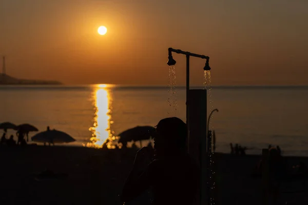 Man silhouette taking shower after swimming at the sea beach. Beautiful view of orange sunset seascape. Summer resort vacation