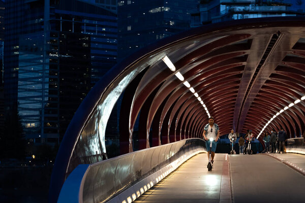 Калгари Альберта Канада, 16 мая 2022: Турист в движении едет на электрическом скутере через мост Мира ночью с пешеходами в популярном районе.