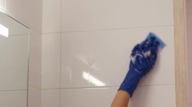 Beyaz tişörtlü bir kadın banyodaki ya da tuvaletteki duvarı mavi temizlik süngeri ve köpüklü deterjan kullanarak yıkıyor. Mavi lastik eldivenli bir el. Duvarda beyaz kiremit