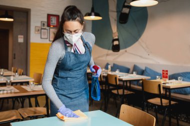 Önlüklü, maskeli ve eldivenli genç garson dezenfektan ve bezle masaları temizliyor.