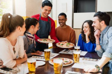 Masada oturan çok ırklı arkadaş grubu garsonun getirdiği pizzalara aç bakıyor..