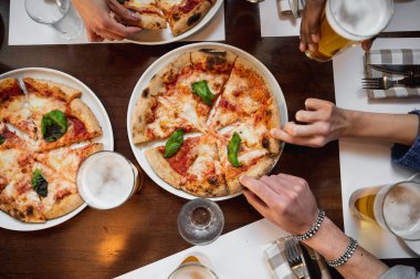 İtalyan Margherita pizzası ve bir bardak birayla en iyi restoran masası. Tanınamayacak kadar çok ırklı insanların elleri dilimlenmiş bir parçayı tutuyor..