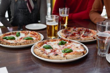 Masanın üzerinde yakın plan bira ve pizza bardakları. Arka planda tanımlanamayan odaklanmış insan grubu.