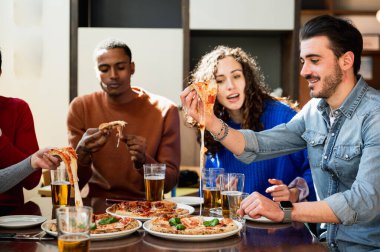 Çok ırklı bir arkadaş grubu masada yemek yiyor. Pizzaların iplikçik peyniri var..