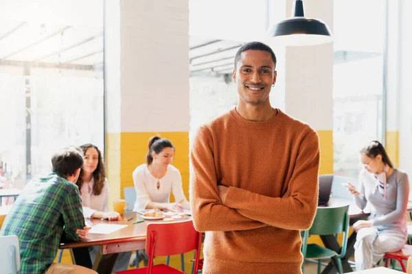Sıradan giyinmiş, kollarını kavuşturmuş, gülümseyen, arka planda oturan bir grup insanla kameraya bakan genç bir Afrikalı..