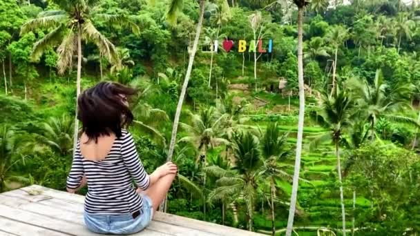 Jong meisje zit en draait langzaam haar hoofd op platform in de buurt van palmbomen en rijst terrassen op Bali. — Stockvideo