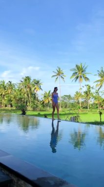 Bali adasındaki pirinç teraslarına ve palmiyelere bakan sonsuz havuzun kenarında dinlenen ve yürüyen güzel kadınlar. Palmiye ağaçları suya yansıyor..