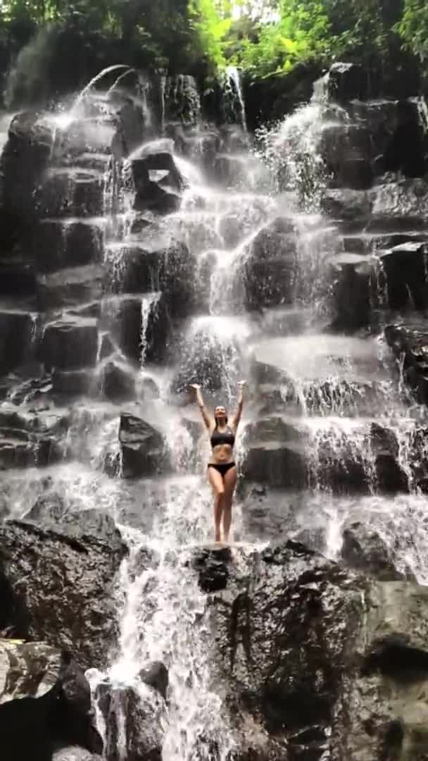 Güzel vücutlu güzel bir kadın şelalenin altındaki bir kayanın üzerinde duruyor Kanto Lampo Bali — Stok video