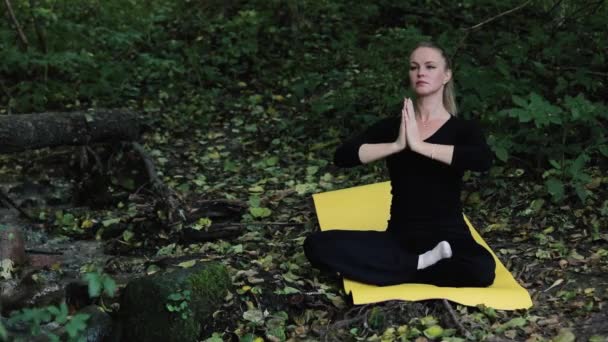 Beautiful middle aged woman practice yoga in autumn forest near river. Female seats on yellow yoga mat on ground in covered by dry leaves woodland and doing exercises. — Stock Video
