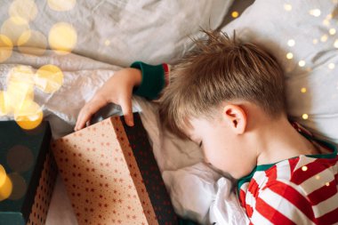 Beyaz yatakta uyuyan çocuk elinde Noel hediyeleri tutuyor..