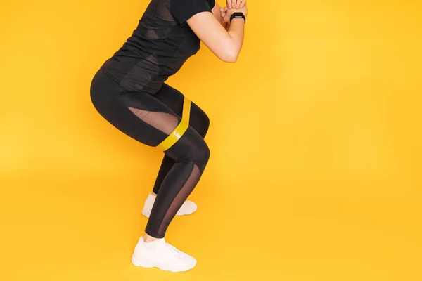 Girl crouches with an elastic band on her legs against yellow background — Stock Photo, Image
