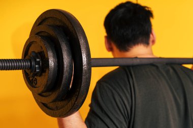 Adam sarı arka planda halteri omuzlarında tutuyor.