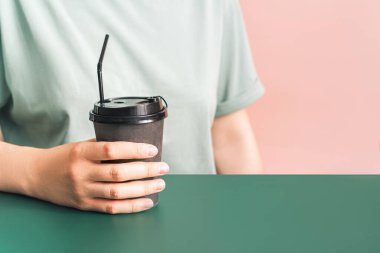 Elinde koyu fincanı olan bir kız pembe arka planda yeşil masada oturuyor.