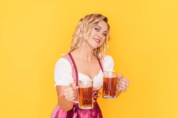 Woman in festive dress holding two mugs with beer in front of yellow background — стоковое фото