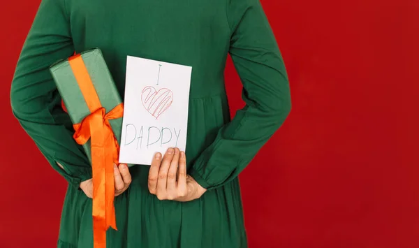 Girl in green dress holds gift and postcard behind her back against red background — Fotografia de Stock