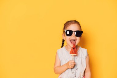 Gözlüklü kız sarı arka planda karpuz şeklinde lolipop yalıyor..