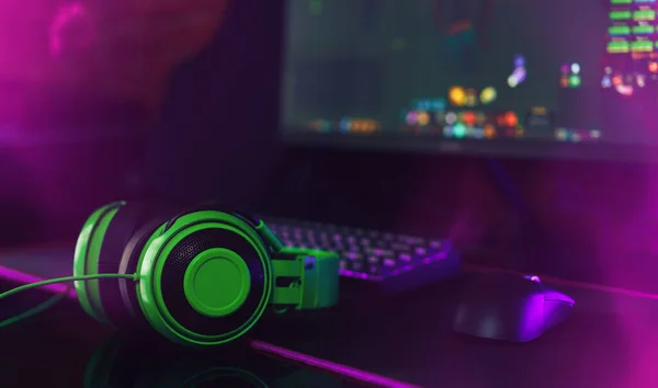 Computer gaming mouse and keyboard with purple backlight next to green headphones opposite the monitor.