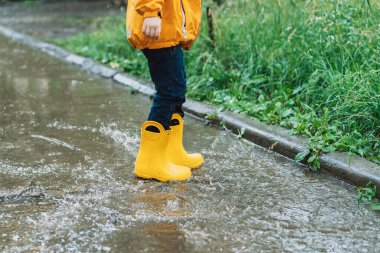 Child in yellow rubber boots jumping through puddles.
