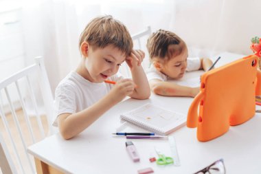 Children draw with felt-tip pens in a notebook and watch a lesson on a tablet.