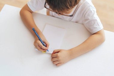 A boy in eyeglasses writes letters in a notebook with a pen.